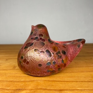 May include: A decorative glass bird sculpture in shades of red, brown, and gold. The art glass bird has a speckled pattern and a textured surface. The bird has a small opening on the side.