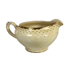 May include: A cream-coloured ceramic creamer featuring a scalloped gold rim and a delicate pattern of small gold dots encircling the upper body. The vessel includes a curved handle on one side and a pouring spout, with a gold band at the base.