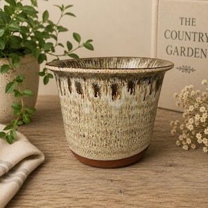 May include: A ceramic planter with a flared rim, featuring a reactive glaze in shades of cream, beige, and dark brown that drips downwards. The base of the planter is unglazed, revealing a natural terracotta-coloured clay finish.