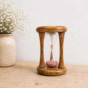 May include: An hourglass with a polished brown wooden frame and clear glass bulbs containing fine pink sand. The hourglass is positioned upright, featuring a circular top and base connected by three vertical wooden pillars, showcasing a smooth finish.