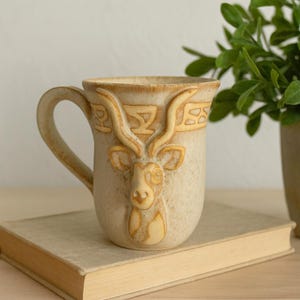 May include: A beige ceramic mug featuring a raised, sculpted relief of an antelope head with long, curved horns. The mug has a speckled finish and a decorative band of geometric patterns encircling the upper rim, complete with a curved handle.
