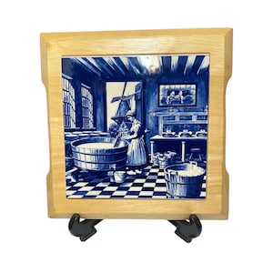 May include: A square tile with a wooden frame, showcasing a blue and white illustration of a Dutch scene. The artwork depicts a person working in a room with a windmill visible through a window. The tile is displayed on a black stand.