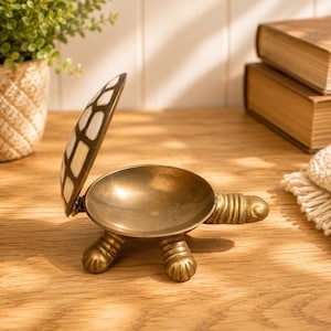 May include: A brass turtle-shaped trinket box with an open lid. The shell lid features a white inlaid geometric pattern. The body of the turtle is made of polished brass with a smooth, circular interior basin and four textured, clawed feet.