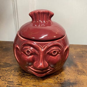 May include: A glossy, dark red ceramic jar with a matching lid. The jar features a moulded, three-dimensional human face on its front, including defined eyes, a nose, and a smiling mouth. The lid has a decorative, ruffled, or pleated top handle.