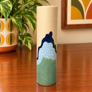 May include: A cylindrical ceramic vase with a matte beige upper section, a central band featuring a dark blue mountain-like shape over a light blue area with small white patterns, and a textured green base with a speckled finish.