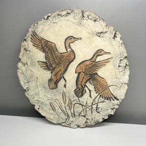 May include: A circular wall plaque featuring a textured, off-white stone-like finish with a relief carving of two brown ducks in flight. The ducks are depicted above a cluster of cattails, with the entire piece showcasing a rugged, uneven edge.