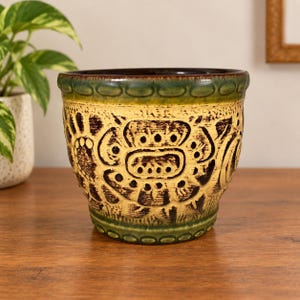 May include: A ceramic plant pot featuring a cream-coloured body with dark brown embossed floral patterns. The rim and base are finished in a deep green glaze with a repeating oval texture. The interior of the pot is a dark brown colour.