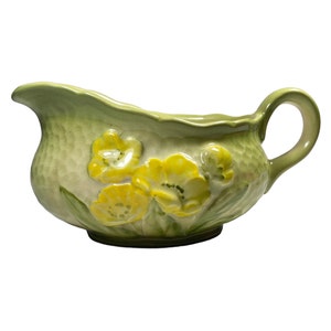 May include: A light green ceramic gravy boat with a textured surface and a handle. The boat features raised yellow floral designs with green leaves. The spout is curved, and the overall design is reminiscent of vintage tableware.