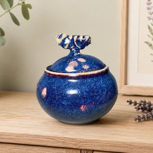 May include: A round ceramic lidded jar featuring a deep blue glaze with speckled white and light pink accents. The lid is topped with a sculptural, textured handle that mimics the appearance of a small, stylised branch or organic knot.