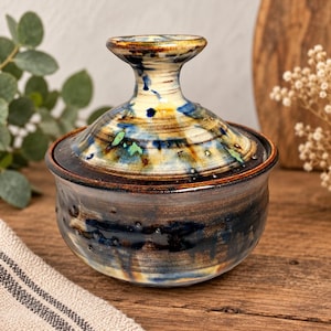 May include: A ceramic lidded jar featuring a rounded base and a matching lid with a prominent knob handle. The surface displays a reactive glaze with blended tones of deep blue, earthy brown, cream, and hints of green in a mottled pattern.