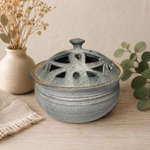 May include: A round, speckled blue-grey ceramic bowl with a matching lid featuring a decorative cut-out pattern. The bowl has a smooth, rounded base and a small knob handle on the centre of the lid, showcasing a textured, mottled glaze finish.