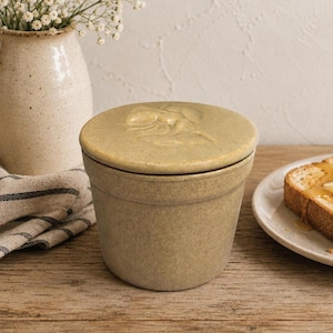 May include: A cylindrical ceramic butter crock with a speckled tan finish and a matching lid featuring a raised, embossed bee design on the top surface. The container has a simple, straight-sided profile and a smooth, matte-textured exterior.