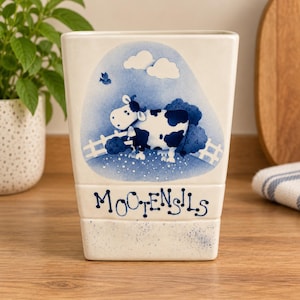 May include: A cream-coloured ceramic utensil holder with a tapered rectangular shape. The front features a blue painted illustration of a cow in a field with clouds and a fence. The text MOOTENSILS is printed in blue below the cow illustration.