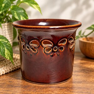 May include: A dark brown glazed ceramic planter featuring a wide, flared rim and a decorative band of cut-out butterfly shapes encircling the upper portion of the vessel. The surface has a glossy, reflective finish with subtle tonal variations.