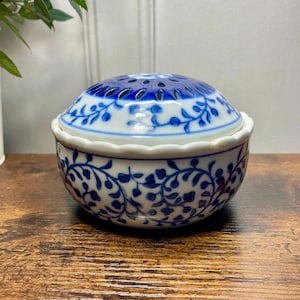 May include: A round white ceramic bowl with a lid featuring a blue floral vine pattern. The lid has a domed shape with a scalloped edge and a series of small circular cutouts at the top. The bowl has a matching blue vine design and a scalloped rim.
