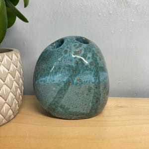 May include: A teal ceramic toothbrush holder with two holes. The holder has a speckled, glossy finish and a rounded, organic shape. The holder is sitting on a wooden surface.