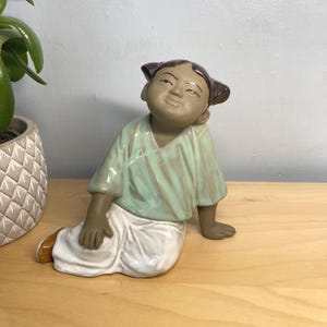 May include: A ceramic figurine of a seated person with a serene expression. The figure wears a light green shirt and white trousers. The figurine is on a wooden surface, with a potted plant visible.