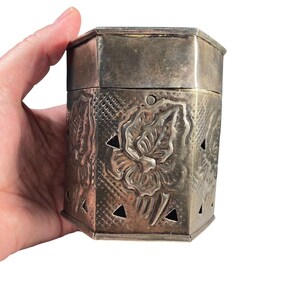 May include: An antique silver-coloured, octagonal box with a hinged lid. The box has embossed floral patterns and triangular cutouts. The box is held in a hand against a white background. The box appears to be made of metal.