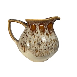 May include: A ceramic jug with a handle and spout. The jug features a speckled brown and cream glaze, with a darker brown rim and handle. The base of the jug is cream-coloured.