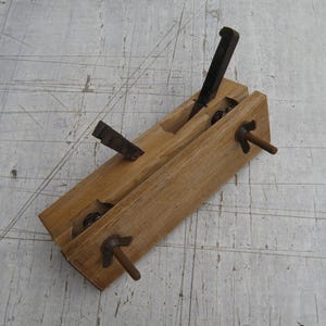 May include: A wooden hand plane tool featuring a light brown rectangular body with two dark metal blades protruding from the top. The tool includes two dark metal wing nuts on threaded rods extending from the side, used for adjusting the blade depth.