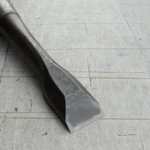 May include: A steel wood chisel featuring a dark, weathered metal handle and a sharp, polished cutting edge. The metal blade displays faint Japanese kanji characters stamped near the neck, indicating the tool's origin and construction.