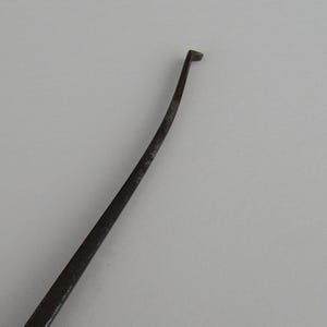 Peut inclure: Un outil en métal foncé et incurvé, avec une pointe plate et rectangulaire. L'outil a un corps fin et allongé qui se rétrécit en pointe. La pointe est légèrement courbée et l'outil semble être vieilli ou utilisé.