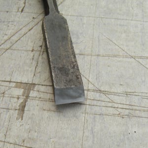 May include: A metal wood chisel featuring a dark, weathered steel shaft and a sharp, polished silver cutting edge. The tool has a rectangular profile with a tapered neck leading toward the handle, showing signs of heavy use and oxidation.