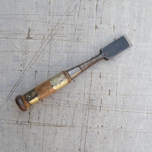 Könnte beinhalten: Ein Holzmeißel mit einer flachen, rechteckigen Metallklinge und einem zylindrischen Holzgriff. Der Griff ist mit einer Metallzwinge nahe der Klinge und einer Metallkappe am Ende verstärkt und weist Gebrauchsspuren sowie eine gealterte Patina auf.
