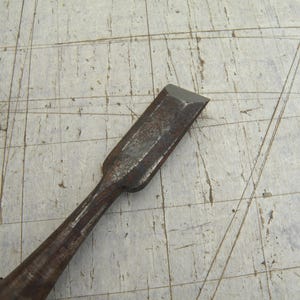 Peut inclure: Un ciseau à bois en métal avec une finition sombre et patinée, doté d'un tranchant biseauté. L'outil présente une tige longue et fine qui s'élargit vers une lame plate, montrant des signes d'oxydation et d'usure sur toute sa surface.