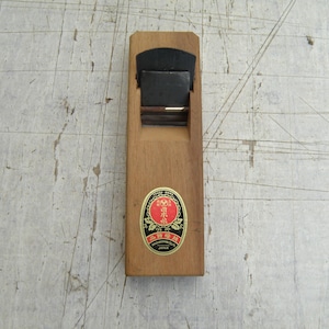 May include: A Japanese wooden hand plane featuring a rectangular light brown wooden body and a dark metal blade set into the top. An oval sticker with a red centre, black border, and gold text is affixed to the front of the wooden block.