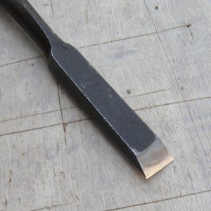 May include: A black metal wood chisel featuring a long, rectangular shaft that tapers into a sharp, polished silver-coloured cutting edge. The tool has a dark, matte finish along the body, contrasting with the bright, metallic bevel at the tip.