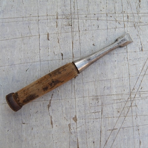 May include: A vintage wood chisel with a metal blade and a wooden handle. The chisel has a worn appearance, suggesting it has been used for woodworking projects. The handle is a light brown color.