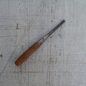 May include: A wood chisel featuring a cylindrical wooden handle and a tapered metal shaft with a flat, sharpened cutting edge. The tool exhibits signs of wear on the metal surface, including discolouration and minor surface imperfections.