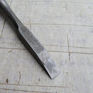 Op de afbeelding: Close-up van een metalen beitel met een platte, afgeschuinde rand. De beitel is donkergrijs van kleur en lijkt goed gebruikt. De achtergrond is een lichtgekleurd, verweerd houten oppervlak.