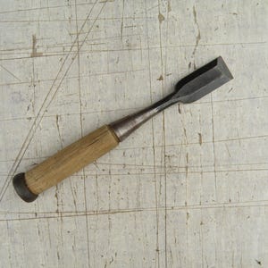 May include: A wood chisel featuring a metallic blade with a beveled edge and a cylindrical wooden handle. The tool has a dark metal ferrule connecting the blade to the handle, and a dark metal cap at the base of the wooden grip for impact resistance.