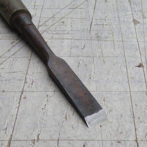 May include: A metal wood chisel featuring a dark, weathered steel blade with a polished, sharp cutting edge and a partially visible wooden handle. The tool exhibits signs of age and use, with a textured, oxidised surface on the metal shaft.