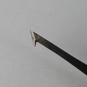 Op de afbeelding: Close-up van een metalen gereedschap met een scherpe, schuine punt. Het gereedschap heeft een donkere, verouderde uitstraling, wat suggereert dat het vintage of veel gebruikt kan zijn. De achtergrond is effen lichtgrijs.