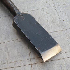 May include: A Japanese wood chisel featuring a dark, forged steel blade with a polished, beveled cutting edge. The metal surface shows a distinct texture from the forging process, and a small, stamped maker's mark is visible near the neck.