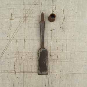 Könnte beinhalten: Ein Vintage-Metallmeißel mit Holzgriff und separater Metallhülse. Der Meißel hat eine rechteckige Klinge und einen konischen Griff. Die Gegenstände liegen auf einer hellen, verwitterten Oberfläche.