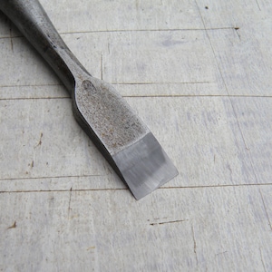 Può includere: Primo piano di uno scalpello in metallo con un bordo piatto e smussato. L'utensile ha un aspetto strutturato e invecchiato, con un manico leggermente curvo. Lo scalpello è progettato per la lavorazione del legno ed è appoggiato su una superficie di legno chiaro.