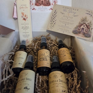 May include: A gift box containing five amber glass spray bottles with beige labels, a bookmark with a pink tassel, and a printed card titled Continue the Story. The labels feature illustrations and text including Cozy Flannel, Pumpkin, and Vanilla Dreams.