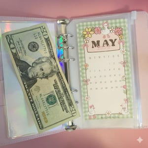 May include: A pink binder with a silver ring mechanism, holding a twenty-dollar bill and a May calendar insert. The calendar has a green gingham border with floral and duck illustrations. The bill is partially visible, showing Andrew Jackson's portrait.