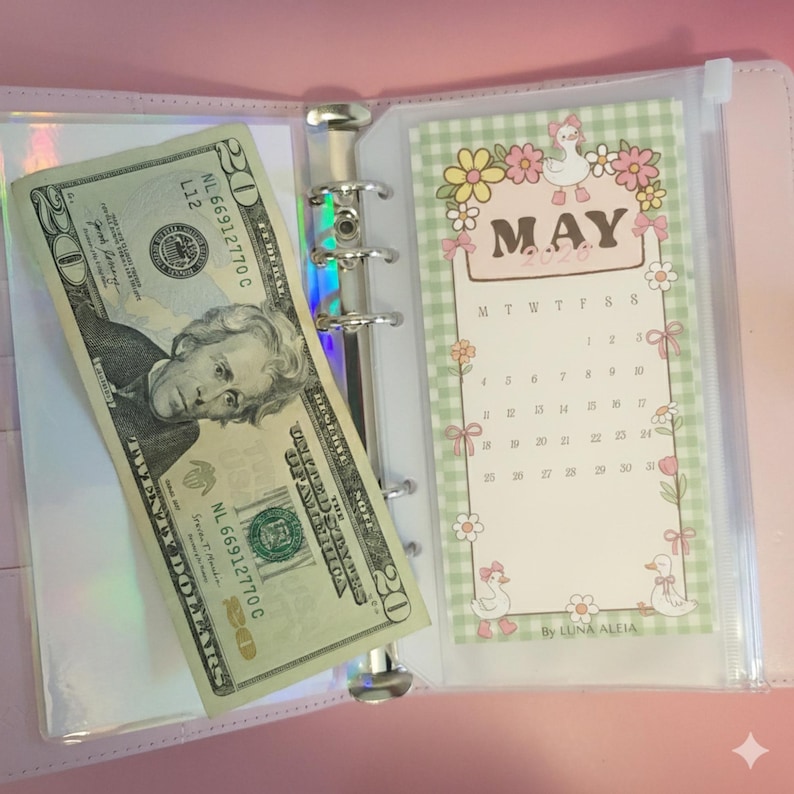 May include: A pink binder with a silver ring mechanism, holding a twenty-dollar bill and a May calendar insert. The calendar has a green gingham border with floral and duck illustrations. The bill is partially visible, showing Andrew Jackson's portrait.