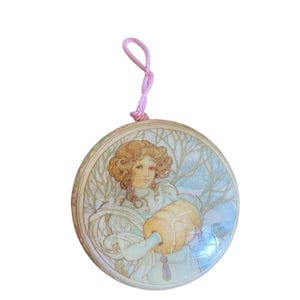 May include: Round ornament featuring a woman with curly hair in a winter scene. The artwork has a vintage style with soft colors and a pink string for hanging. The woman is holding a bundle.