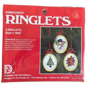 May include: A red package of Embroidery Ringlets, style 9001, containing three 2x2.75 inch oval goldtone frames, fabric, needle, and DMC floss. The packaging displays three finished oval ornaments featuring a snowman, a tree, and a poinsettia.