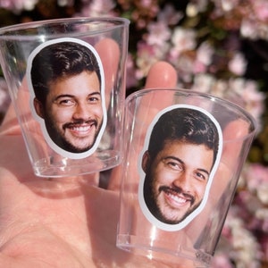 May include: Two clear plastic shot glasses with a printed image of a person's face on each. The person has brown hair and a beard.
