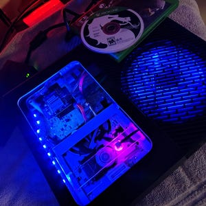 May include: Black Xbox One console with blue internal lighting and a game disc. The console has a transparent section revealing internal components. A power supply is visible to the left. The game disc is in a green case.