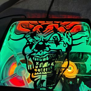 May include: A black and green illuminated sign featuring a menacing clown face. The clown's face is a silhouette with a detailed skull design. The sign has a red and orange top and a green border, with internal lighting.