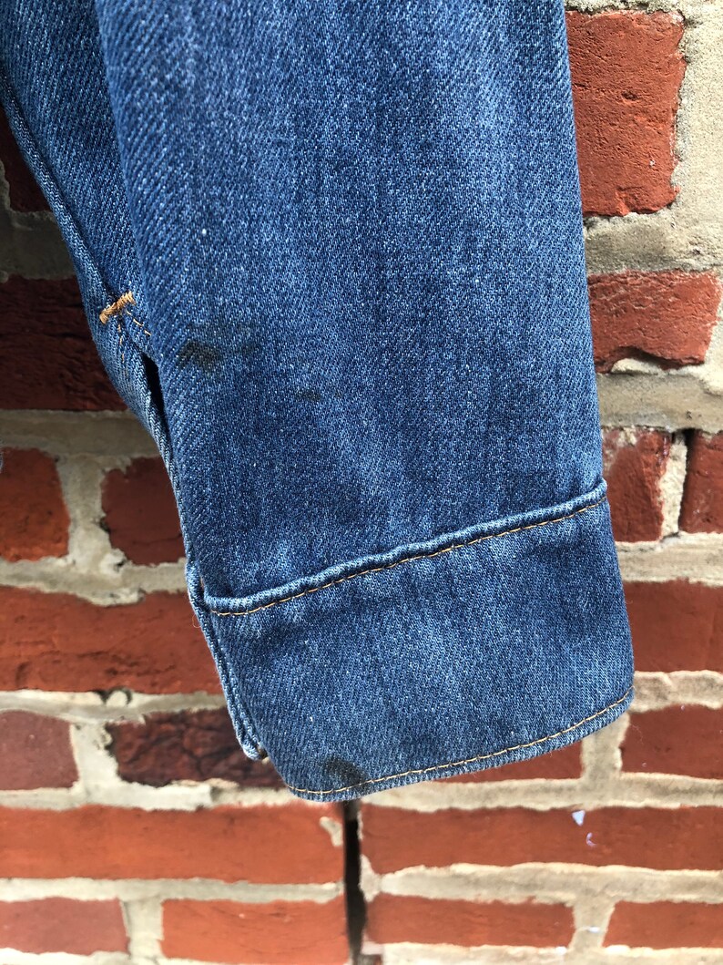 May include: Close-up of the cuff of a blue denim jacket with a yellow stitch detail.