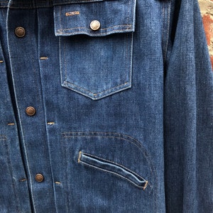 May include: A blue denim jacket with a button closure, two chest pockets, and a single pocket on the left sleeve. The jacket has a worn look and is made of a thick, durable fabric.